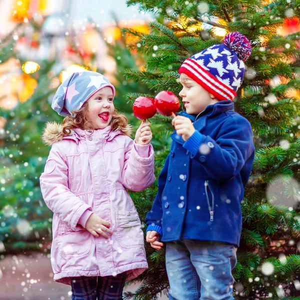Dvě malé usmívající se děti, chlapec a dívka jíst slazené apple na vánoční trh. Šťastné děti v zimě módní oblečení s osvětlením na pozadí. Srandovní sestra a bratr. — Stock fotografie