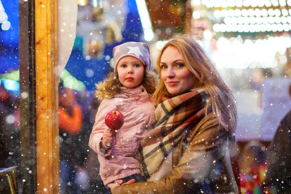Mujer hermosa y niña pequeña comiendo manzana azucarada cristalizada en el mercado de Navidad alemán. Familia feliz, madre e hija en ropa de invierno con luces en el fondo. Tradición, vacaciones —  Fotos de Stock