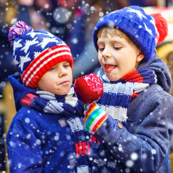 Dva malí kluci kluk, roztomilý sourozenci jíst cukr apple poblíž sweet stojan s ořechy a perníčky. Šťastné děti na vánoční trh v Německu. Tradiční odpočinek na Vánoce — Stock fotografie