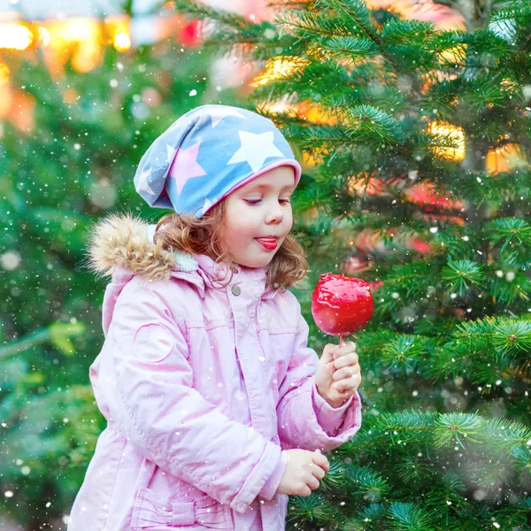 Cute little kid girl eating sugared apple on Christmas market. Happy child in winter fashion clothes with lights on background — Stock Photo, Image