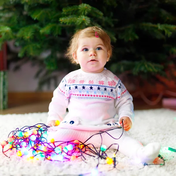 Adorable baby girl holding colorful lights garland in cute hands. Little child in festive clothes decorating Christmas tree with family. First celebration of traditional feast — Stock Photo, Image