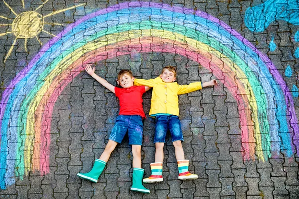 Twee schooljongens die plezier hebben met regenboogtekeningen met kleurrijk krijt op asfalt. Broers en zussen, tweelingen en beste vrienden in rubber laarzen schilderen op de grond samen spelen. — Stockfoto