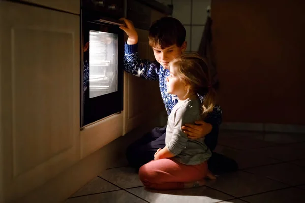 En liten barnepike og en søt gutt som baker muffins og småkaker på kjøkkenet. To barn, søsken, bror og søster som sitter ved siden av ofen og venter. På thanksgiving eller helligdag . – stockfoto