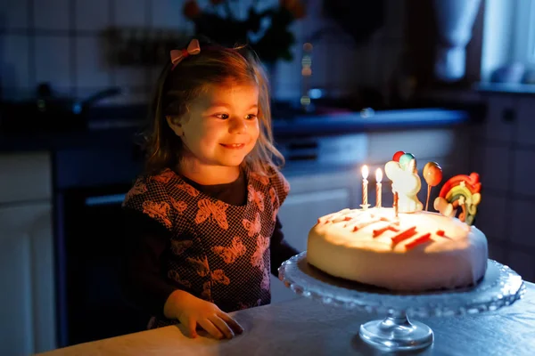 Adorable niñita celebrando su tercer cumpleaños. Bebé niño pequeño con pastel de unicornio casero, interior. Feliz niño sano es sorprendido acerca de chispa de fuegos artificiales y soplar velas en la torta — Foto de Stock