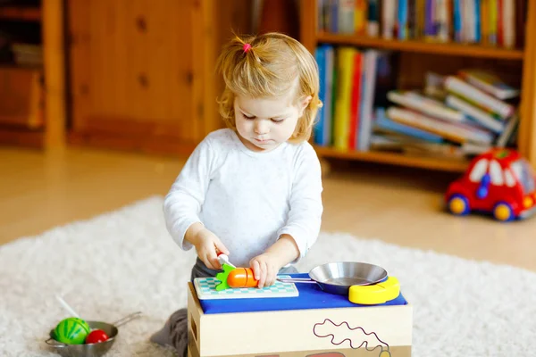 Schattig klein peutermeisje dat thuis speelt met eco houten speelgoed. Gelukkig gezond opgewonden kind snijden groenten en fruit met speelgoed mes. Baby meisje hebben plezier met rol keuken en koken spel. — Stockfoto