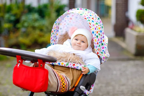 Bonito pequena menina bonita sentada no carrinho de bebê ou carrinho no dia de outono. Criança sã feliz que vai para um passeio no ar puro na roupa quente. Bebê com roupas coloridas e chapéu com bobbles — Fotografia de Stock