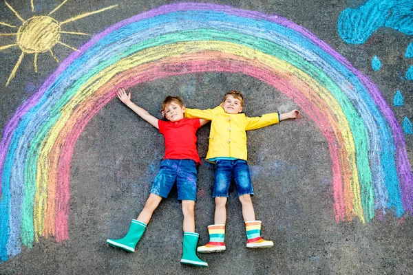 Dois meninos da escola que se divertem com com desenho do arco-íris com giz colorido no asfalto. Irmãos, gêmeos e melhores amigos em botas de borracha pintando no chão brincando juntos . — Fotografia de Stock