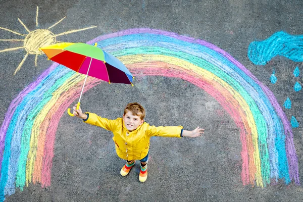 Ragazzo felice in stivali di gomma con sole arcobaleno e nuvole con gocce di pioggia dipinte con gessetti colorati a terra o asfalto in estate. Tempo libero creativo per bambini all'aperto in estate — Foto Stock