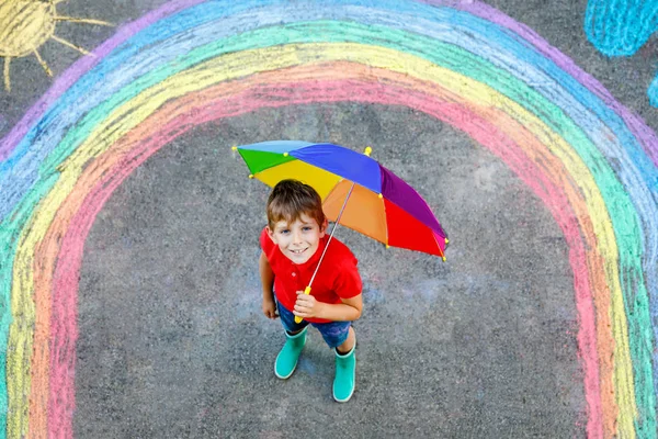 Ragazzo felice in stivali di gomma con sole arcobaleno e nuvole con gocce di pioggia dipinte con gessetti colorati a terra o asfalto in estate. Tempo libero creativo per bambini all'aperto in estate — Foto Stock