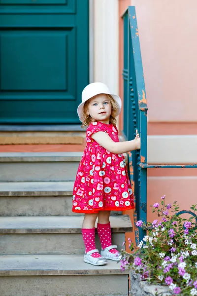 Portret van prachtige kleine gorgeus mooie peuter meisje in roze zomer look kleding, mode jurk, knie sokken en hoed. Gelukkig gezond baby kind poseren voor de voorkant van kleurrijke huis. — Stockfoto