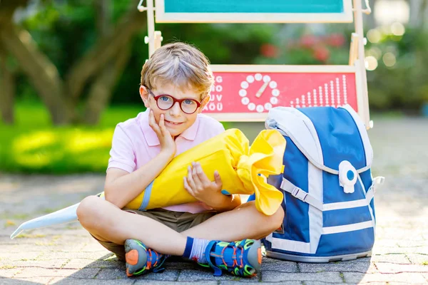 Gelukkige kleine jongen met een bril aan zijn bureau en rugzak of tas. Schoolkind met traditionele Duitse schooltas kegel genaamd Schultuete op zijn eerste dag naar school — Stockfoto