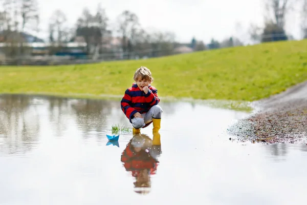 Happy little kid boy in yellow rain boots playing with paper ship boat by huge puddle on spring or autumn day — Stock Photo, Image