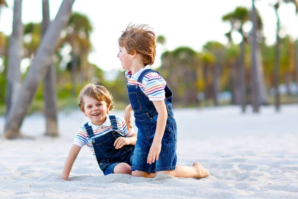 İki küçük çocuk tropikal plajda eğleniyor, mutlu arkadaşlar oynuyor, arkadaşlık kavramı. Kardeşler, ikizler kavga ediyor, koşuyor ve ailede zıplıyor. Arka planda palmiye ağaçları var.. — Stok fotoğraf
