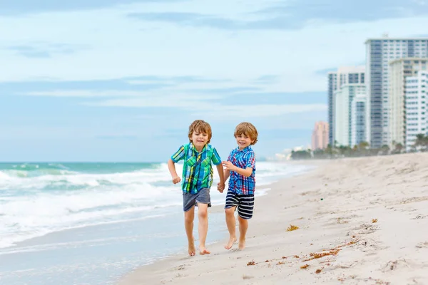 Dwóch szczęśliwych chłopców biegających po plaży oceanu. Śmieszne słodkie dzieci, rodzeństwo i najlepszych przyjaciół dokonywania wakacji i ciesząc się latem w burzliwy wietrzny dzień. Zdrowe dzieci na plaży w Miami, USA — Zdjęcie stockowe