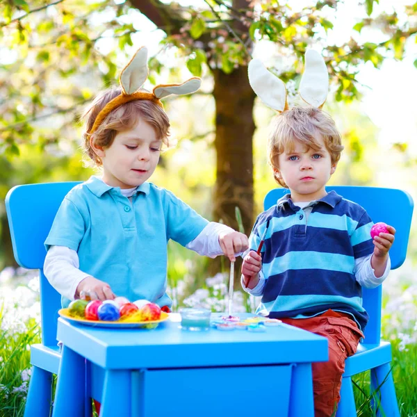 Two cute little kid boys wearing Easter bunny ears, painting colorful eggs and having fun outdoors. Family, siblings, brothers, twins celebrating religious holiday. Best friends coloring eggs. — Stock Photo, Image