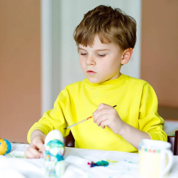 Piccolo bambino in età prescolare biondo che colora uova per vacanza di Easter in cucina interna, all'interno. Bambino divertendosi con la pittura colori e festeggiando festa — Foto Stock