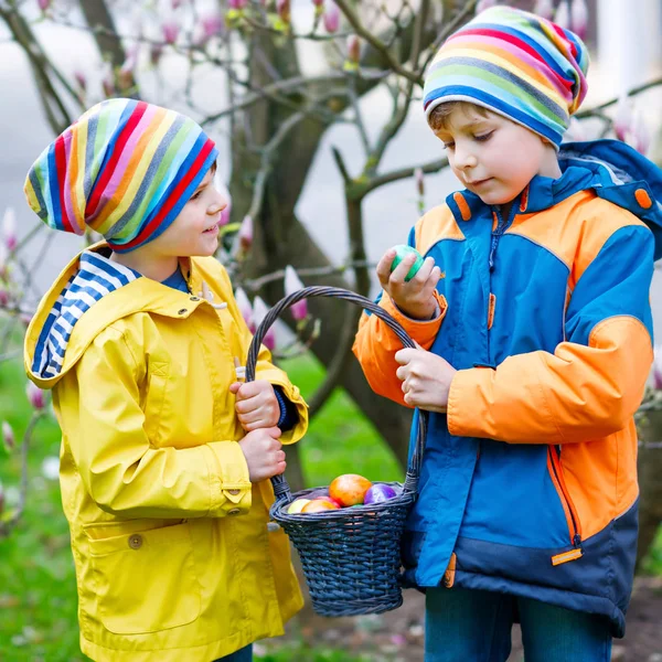 Doi băieți mici și prieteni care fac vânătoare tradițională de ouă de Paști în grădina de primăvară, în aer liber. Frații se distrează cu găsirea ouălor colorate. Într-o zi rece. Vechea tradiție creștină și catolică . — Fotografie, imagine de stoc