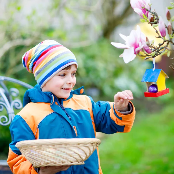 Cute adorable little kid boy making an egg hunt on Easter. Happy child searching and finding colorful eggs in domestic garden. Boy in spring clothes on cold day. Old christian and catholoc tradition. — Stock Photo, Image