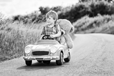 Küçük anaokulu çocuğu büyük oyuncak araba kullanıyor ve dışarıda pelüş oyuncak ayısıyla oynarken eğleniyor. Çocuk, doğada ılık yaz gününün tadını çıkarıyor..