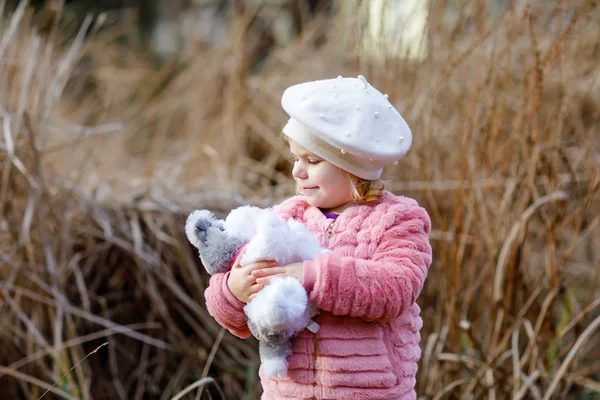 Retrato al aire libre de pequeña niña linda en abrigo rosa y sombrero de moda blanco barret jugando con peluche suave juguete para perros. Niño feliz y saludable caminando en el parque en un día frío . —  Fotos de Stock