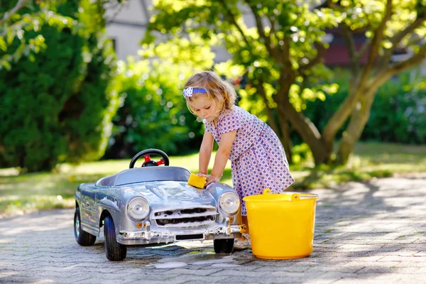 Carino splendida bambina lavare grande vecchia macchina giocattolo nel giardino estivo, all'aperto. Buon bambino sano pulizia auto con acqua e sapone, divertirsi con spruzzi e giocare con la spugna. — Foto Stock