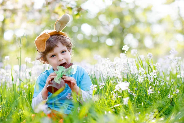 Netter kleiner Junge mit Osterhasenohren feiert traditionelles Fest. Glückliches Kind, das an einem warmen, sonnigen Tag Schokohasen-Fugure isst. Familie, Urlaub, Frühlingskonzept. Kleinkind sitzt zwischen Blumen — Stockfoto