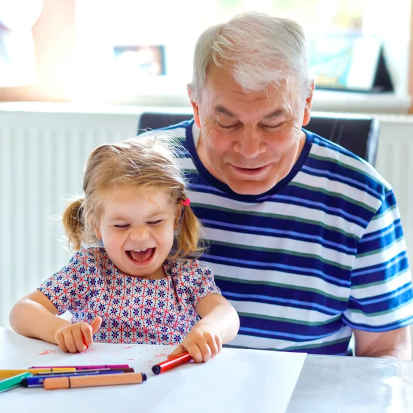 Schattig klein baby peuter meisje en knappe senior grootvader schilderij met kleurrijke potloden thuis. Kleinkind en man hebben plezier samen. Familie en generatie in liefde — Stockfoto