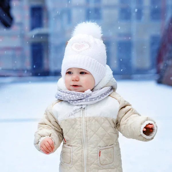 Schattig klein meisje dat 's winters de eerste stappen buiten zet. Leuke peuter die leert lopen. Kind heeft plezier op koude sneeuwdag. Het dragen van warme baby roze kleren en hoed met bobbels. — Stockfoto