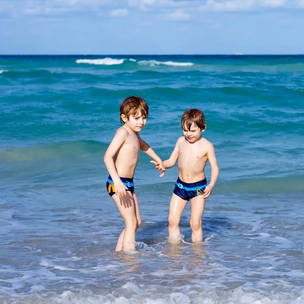 Dos niños felices corriendo en la playa del océano. Divertidos niños lindos, hermanos, gemelos y mejores amigos haciendo vacaciones y disfrutando del día de verano. Divertirse con saltar en la ola. — Foto de Stock