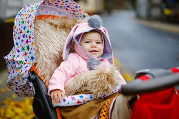 Schattig klein mooi babymeisje zittend in de kinderwagen of kinderwagen op herfstdag. Gelukkig lachend kind in warme kleren, mode stijlvolle roze babyjas met konijnenoren. Baby gaat wandelen met ouders. — Stockfoto