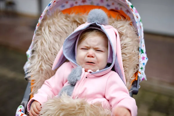 Szomorú sír kis gyönyörű kislány ül a babakocsit vagy babakocsi az őszi napon. Boldogtalan fáradt és kimerült gyermek meleg ruhákat, divat stílusos rózsaszín baba kabát nyuszi füle. — Stock Fotó