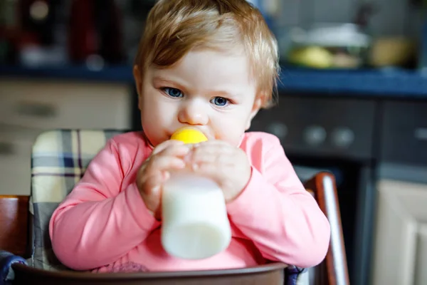 Linda menina adorável segurando mamadeira e bebendo leite fórmula. Primeira comida para bebés. Criança recém-nascida, sentada na cadeira da cozinha doméstica. Bebês saudáveis e conceito de alimentação por mamadeira — Fotografia de Stock