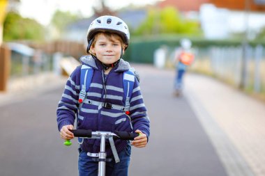 Kasklı aktif okul çocuğu güneşli bir günde şehirde scooterıyla geziyor. Renkli elbiseli mutlu çocuk okula giderken bisiklet sürüyor..