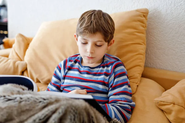 Leuke blonde kleine jongen die tijdschrift leest of boek in de huiskamer. Opgewonden kind dat luid leest, zittend op de bank. Schoolkind, familie, onderwijs. — Stockfoto