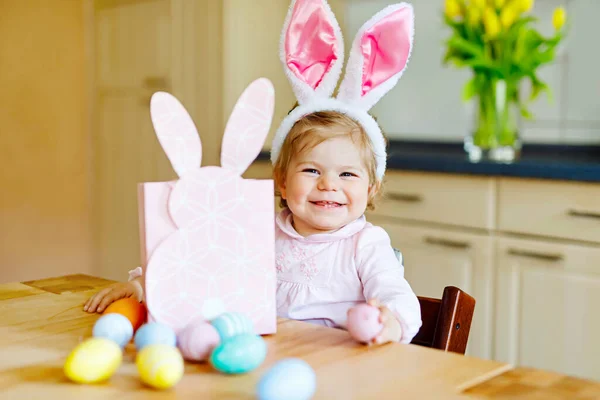 Schattig klein peuter meisje dragen Easter bunny oren spelen met gekleurde pastel eieren. Gelukkig kind uitpakken babygiften. Schattig kind in roze kleding genieten van vakantie — Stockfoto