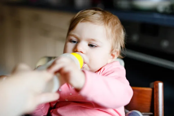 Χαριτωμένο αξιολάτρευτο κοριτσάκι κρατώντας μπουκάλι νοσηλευτικής και πίνοντας γάλα φόρμουλα. Το πρώτο φαγητό για μωρά. Νεογέννητο παιδί, κάθεται στην καρέκλα της οικιακής κουζίνας. Υγιή μωρά και έννοια της διατροφής με φιάλες — Φωτογραφία Αρχείου