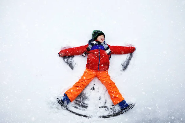 Aranyos kisfiú, színes téli ruhákban hóangyalt csinál, és a hóra fekszik. Aktív szabadtéri kikapcsolódás gyerekekkel télen. Boldog egészséges gyermek szórakozás és nevetés a szabadban — Stock Fotó
