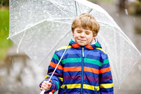 寒い日に傘を持って学校に行く途中の美しい小さな男の子は、雨や雪の中を歩いています。カラフルなファッションで幸せと喜びの子供カジュアル服. — ストック写真