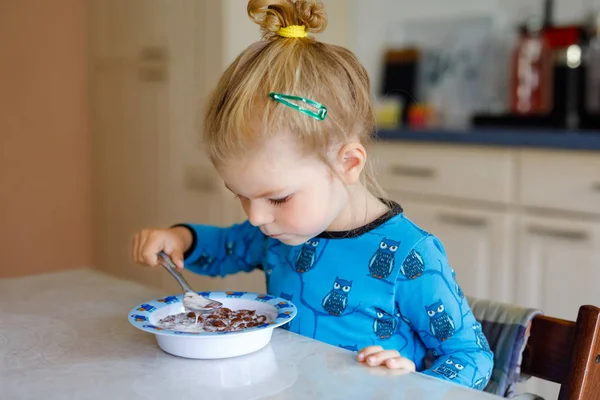 Underbara lilla småbarn flicka äter hälsosamma spannmål med mjölk till frukost. Söt glad baby barn i färgglada kläder sitter i köket och har kul med att förbereda havre, spannmål. Inomhus hemma — Stockfoto