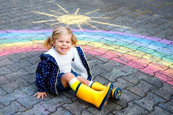 Pequeña niña feliz en botas de goma con sol de arco iris y nubes con lluvia pintadas con tiza de colores en el suelo o asfalto en verano. Lindo niño divirtiéndose. ocio creativo —  Fotos de Stock