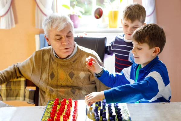 Dědeček a dva kluci si spolu hrají na strategický stůl. Starší muž se baví se svými vnoučaty. Sourozenci děti těší rodinný čas s dědečkem — Stock fotografie