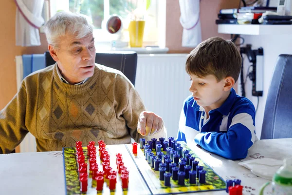 Дедушка и школьник вместе играют в стратегические настольные игры. Старший веселится со своим внуком. Ребенок наслаждается семейным временем с дедушкой — стоковое фото