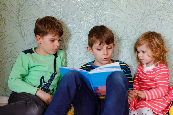 Two school kids boys and little toddler girl reading together book. Older brother holding book and read aloud for his sibling. Family spending time together. Three children. — Stock Photo, Image