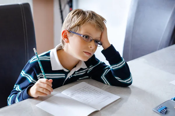 Задумчивый маленький мальчик в очках дома делает домашнее задание, пишет текст красочными ручками. Маленький ребенок занимается спортом в помещении. Начальная школа и образование — стоковое фото