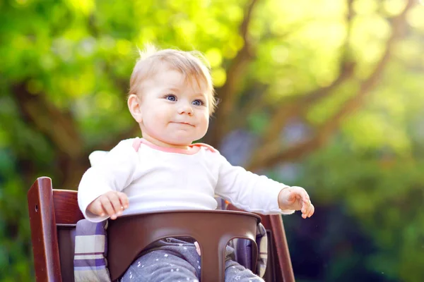 Preciosa adorable niña sentada en la silla alta al aire libre. Beatuiful niño de 6 meses en el jardín en casa, jugando en el día soleado cálido. Bebé sano sonriendo y riendo . — Foto de Stock