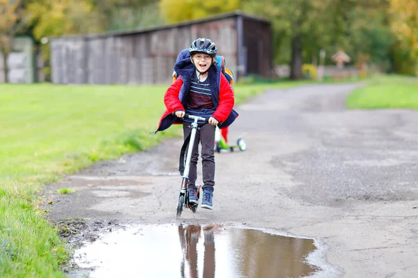 通学途中や通学途中のプッシュスクーターに乗ってる可愛い小学生の男の子。雨の水たまりを通って運転7年の学校の少年。カラフルなファッションの服とヘルメットで面白い幸せな子供. — ストック写真