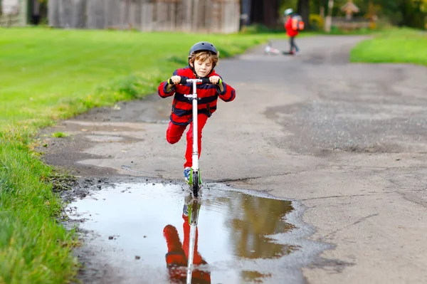 Netter kleiner Schuljunge, der auf seinem Roller auf dem Weg zur oder von der Schule fährt. Schulkind von 7 Jahren fährt durch Regenpfütze. Lustiges fröhliches Kind in bunten Kleidern und mit Helm. — Stockfoto