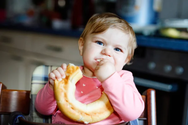 Linda niñita comiendo pan. Adorable niño comiendo por primera vez pedazo de pretzel o croissant. Niño feliz sonriente saludable. Vienen los primeros dientes. En casa dentro de la cocina . —  Fotos de Stock