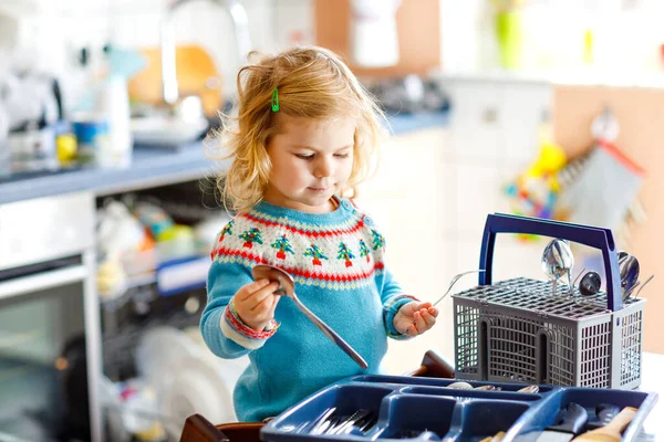 Aranyos kislány segít a konyhában mosogatógéppel. Boldog egészséges szőke gyerek válogató kések, villák, kanalakat, evőeszközök. Baby szórakozás segítésével házimunka anya és apa. — Stock Fotó