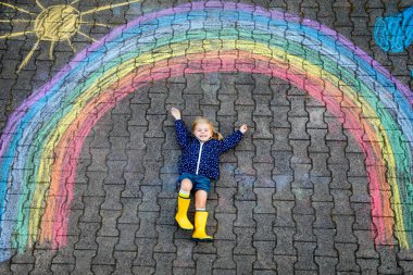 Happy little toddler girl in rubber boots with rainbow sun and clouds with rain painted with colorful chalks on ground or asphalt in summer. Cute child having fun. creative leisure clipart